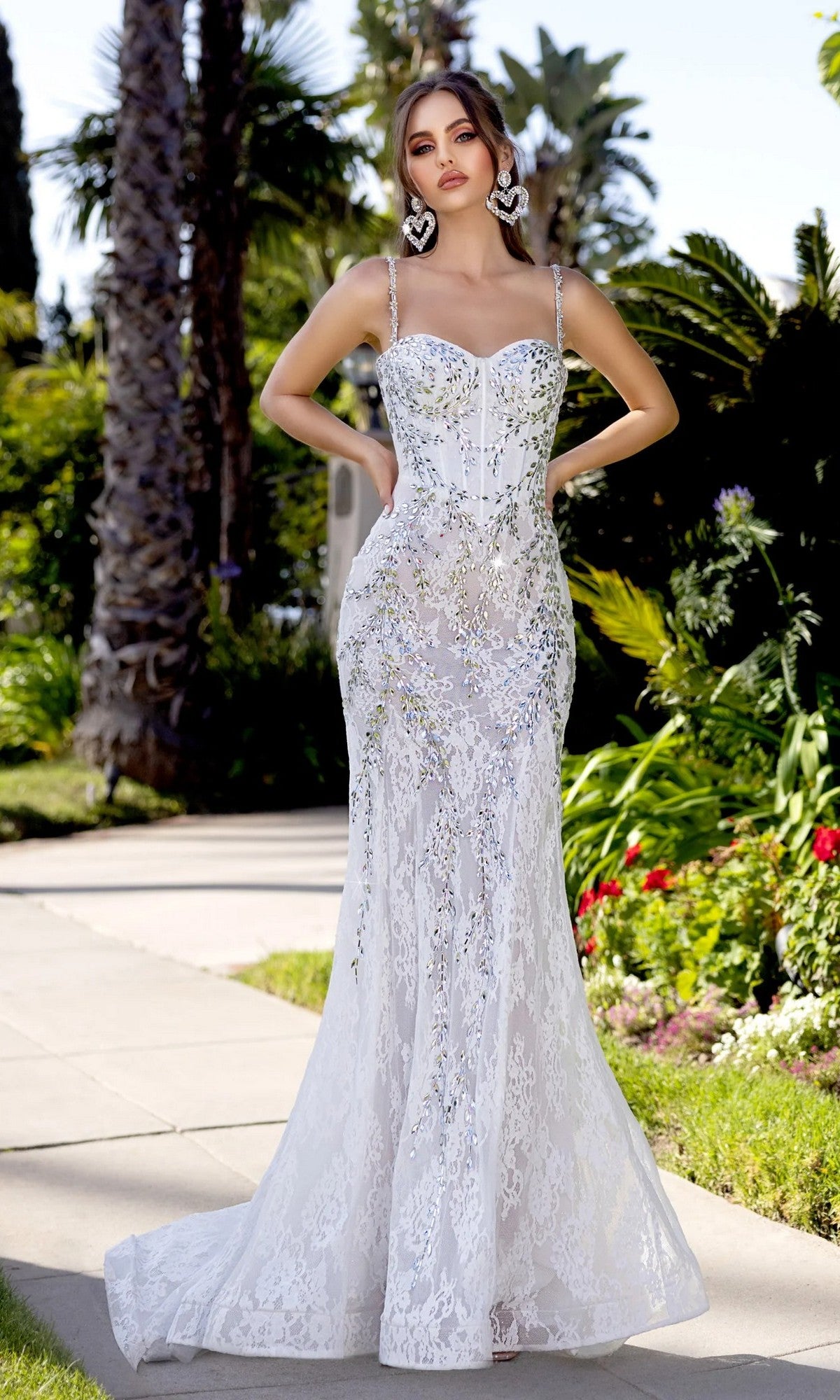 A woman stands outdoors among greenery and palm trees in the Portia and Scarlett PS25150 long prom dress, featuring a fitted ivory/silver lace design with thin straps and silver crystal beading.